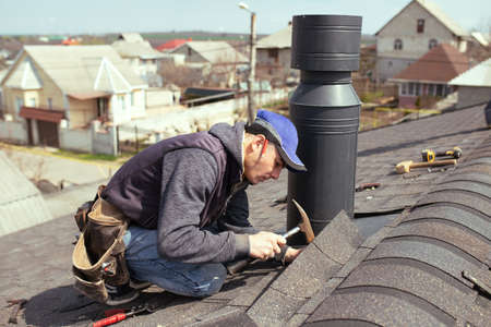 a professional master roofer with hammer repairs the roof. Repair Asphalt Shingles or Bitumen Tiles on the Rooftop Outdoorの写真素材