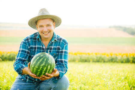 the portrait of a smiling farmer holds a watermelon in his hand. the man looking at the camera is wearing a hat and is in the agricultural fieldの写真素材