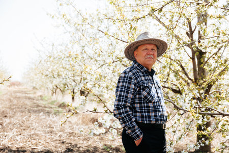 old farmer with hat puts his hands in your pocket. against the background of the flowering orchard, during the springの写真素材