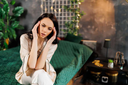 Attractive caucasian brunette woman dressed in pijamas sitting on bed in bedroom and having headache. feeling sad and stressed at home. Disappointed and desperate girlの写真素材