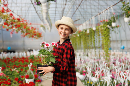 preteen kid happy girl with hat and a flower pot in her hand. smiling child looking at camera is in the greenhouse with many flowers, Copy spaceの写真素材