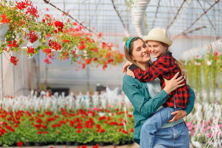 a happy child in the arms of his mother, a family of gardeners who are in the greenhouse. the mother hugs her daughter tightly. smiling mom and little kid, copy spaceの写真素材