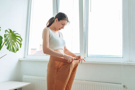 Slim surprised happy woman body in big pants. slender figure tries on big pants and shows how to lose weight. A slim girl in too big pants showing how she was losing, real people, at homeの写真素材