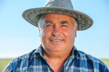 Glad elderly male worker in hat looking at camera while standing in agricultural plantation on sunny summer day in countryside, Content mature farmer in countrysideの写真素材