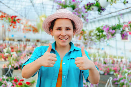 Pretty women stands in a greenhouse with flowers, smiles broadly, looks at the camera and shows thump up. The girl has a hat on her head and a good mood. Around the flowers in pots. Copy spaceの写真素材