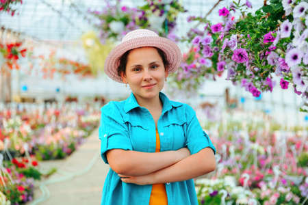 Front view of a young agronomist girl in a greenhouse, the woman crosses her arms over her chest and smiles playfully. The agronomist girl stands in a hat and shirt among the flowers.の写真素材