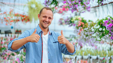 A happy young guy stands in greenhouse with flowers and smiles broadly, looking at the camera, shows thump up. The man man wearing a plaid shirt and has good mood. Copy spaceの写真素材