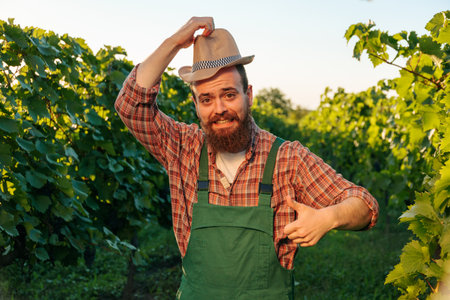 Front view looking at camera green vineyard handsome young bearded winemaking farmer man smiling with one hand greeting holding his hat, the other thumb up. Winemaker happy that harvest was a success.の写真素材