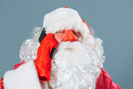 Looking into the camera on a blue background close up front view portrait of Santa Claus speaking on the smartphone. Father Frost decides important matters on the eve of Christmas and New Year.の写真素材