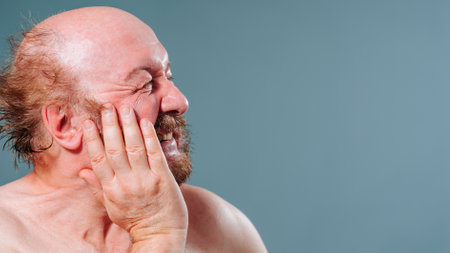 Side view of an elderly man holding his cheek, toothache, mustache, beard, wrinkled face, grimacing in pain, naked. Copy space. Studio isolated blue background.の写真素材