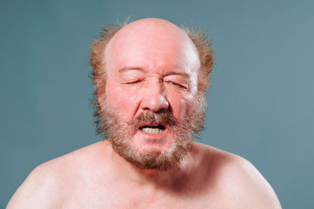 Front view portrait of an elderly man with a beard and mustache wrinkled, expression of pain, sadness, sadness on his face, bare shoulders, open mouth, teeth visible. Studio isolated blue background.の写真素材