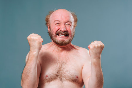Portrait elderly senior man rejoices makes faces against background blue studio his hands are clenched into fists in front of him, gritted teeth in smile. Studio isolated blue background. Copy space.の写真素材