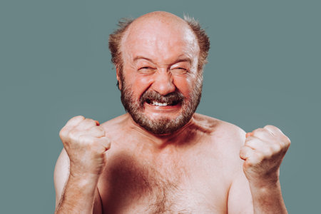 Portrait of a man aged front view looking at camera clenched teeth holding his hands in front of him in fists. An elderly man is bald with a beard and mustache. Studio isolated blue background.の写真素材