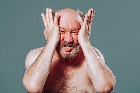 Front view an aged man looking at camera, holds his hands on face with palms to himself, twists and squeezes face from strong different emotions. Studio isolated blue background.の写真素材