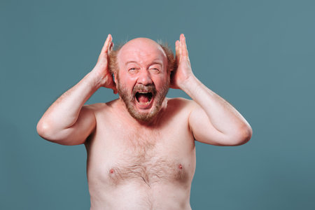 An aged man desperately with all his soul shows his emotions expression on his face hands at head. Front view looking at camera naked torso beard mustache bald. Studio isolated blue background.の写真素材