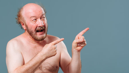 Front view an elderly man stands from the left looking to the right pointing there with the index fingers of both hands. An older man is fooling around and laughing. Studio isolated blue background.の写真素材