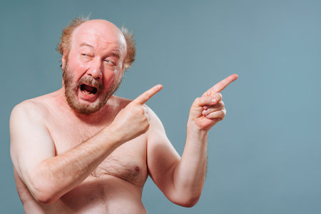 Front view elderly man stand from left looking to left pointing right index fingers of both hands. An older man is fooling around and laughing. Studio isolated blue background. Copy space.の写真素材