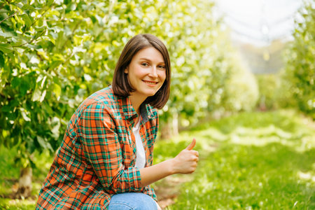 Young woman farmer agronomist gardener is squatting in an apple orchard and smiling showing thumb up. An adult girl is happy that she is doing her favorite work being in nature among fruit trees.の写真素材