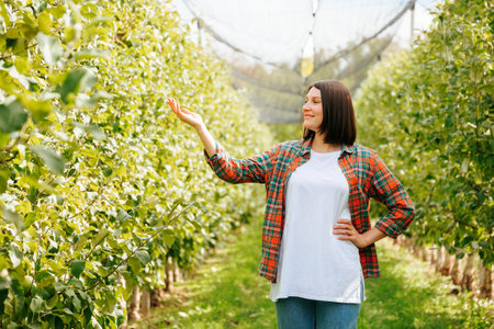 Front view copy space looking away young woman worker smile happy stand in orchard one hand up. Pretty adult girl agronomy in the garden with green trees is glad. Short haircut, brunette hair.の写真素材