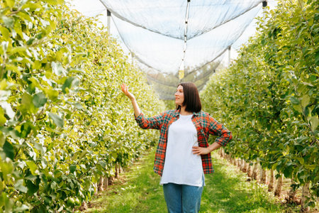 Front view copy space young adult girl woman in orchard one hand up second on hips smile slightly. Youth farmer worker stand in apple garden looking away showing something in checkered shirt.の写真素材