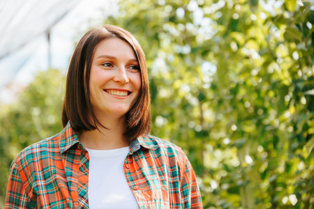 Front view portrait beautiful young woman farmer standing in the garden smiling beautiful gentle happy smile. Looking away. Female has short brunette hair casual wear is white t shirt. Copy space.の写真素材