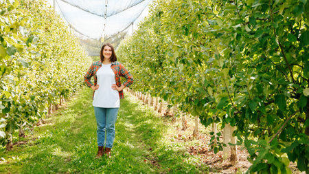 Front view full length pretty woman farmer worker stand in apple orchard smiling lovely arms hips. Female worker agronomy is wearing in casual closes looking at camera, woman brunette, small business.の写真素材