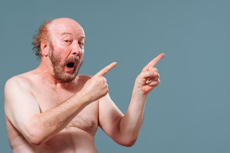 Front view senior man stand left looking at camera pointing index fingers right mouth say wow. Elderly man is fooling around laugh. Studio isolated blue background. Copy space.の写真素材