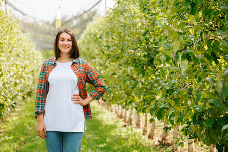 Front view looking at camera charming smile farmer woman short haircut stand in an apple orchard. A cute adult girl farmer beautifully holds her hand on her belt and smiles slightly. Copy space.の写真素材