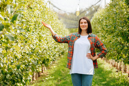 Young woman farmer front view copy space one hand raised up standing in a garden with trees smile. Beautiful adult girl in a white t shirt and jeans. Looking at camera outside photography.の写真素材