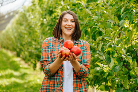 Front view young beautiful woman farmer orchard worker harvested and holding three apples in her hands. The harvest was rich. An adult girl with a short haircut in a plaid shirt.の写真素材