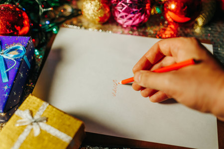 Unrecognizable top view hand with red pencil writes something on paper. Nearby are blue and yellow boxes with gifts. Selective focus, twinkling lights.の写真素材