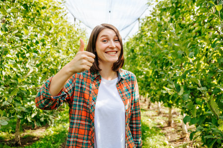 Thumb up with one hand wide smile young woman farmer looking at camera. Copy space in an apple orchard. She is wearing a plaid shirt and a white t shirt. Front view.の写真素材