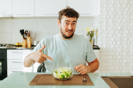 A young dieting man points his index finger at a bowl of salad in front of him, asking Is this supposed to be eaten Green salad healthy food concept. Sits in his kitchen. Looking at camera.の写真素材