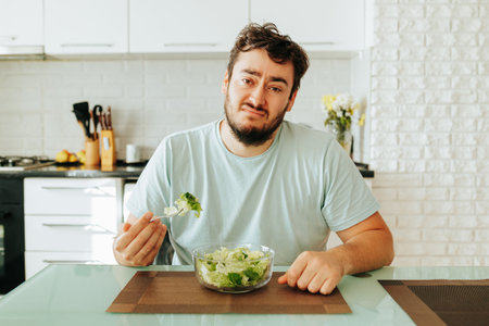 Tired of the diet, dissatisfied with food, a young man sits at a table holding a fork with salad in his hands. He does not like veganism and vegetarianism. Stop diet.の写真素材