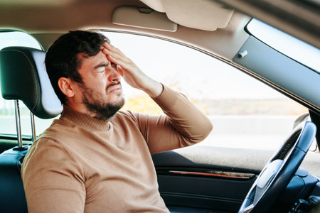 Suffering guy young man is in the car and holds his hand on his head writhing in pain. The forced stop delays the case. A common problem for drivers. Better stop.の写真素材