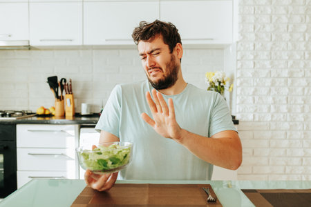 A young man, grimacing, looks towards the salad, how you don t want to eat it. Located in the kitchen. Filming inside. Stop diet. Front view.の写真素材