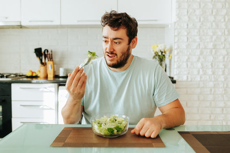 Distraught with hunger, a young guy doomily eats a green salad. Delicious salad and healthy vegetables. Healthy food is boring. Stop diet.の写真素材