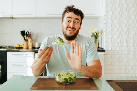 A young guy in a hysterical state renounces vegetables and greens, showing stop with his hand. Front view guy at home in the kitchen. Healthy food. Stop diet.の写真素材