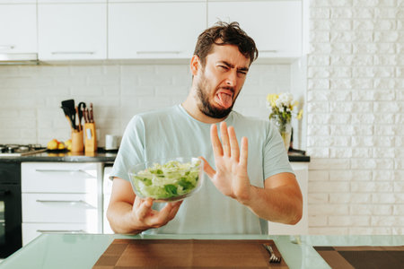 Young man guy is fed up healthy food in the form of greens and vegetables, grimaces at the sight of a full bowl of healthy food.の写真素材