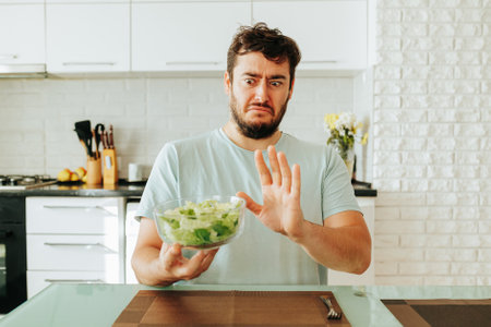 Stop diet guy says with all his appearance grimaces hold bowl of salad in his hand. He looks at dinner with wide eyes and does not want to eat it. Front view. Young man follows a diet without desire.の写真素材