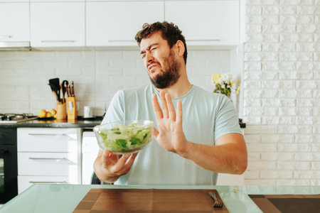A young guy, a man, takes care of his health and is on a diet, grimaces with displeasure and pushes away a bowl of salad. Good and healthy food is the key to health. Front view. Stop diet.の写真素材