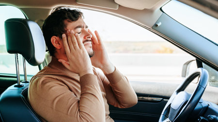 A grimace of pain on the face of a young male driver sitting at the wheel of a car, hands on his temples. He closed his eyes, held his head in his hands, let go of the steering wheel.の写真素材