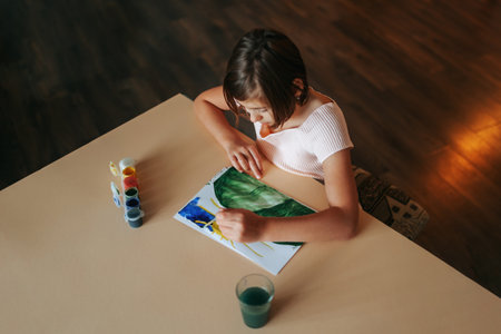 Top view of a nice serious child girl sitting at a desk, drawing something in an album with left hand, looking on album. Pretty kid doing what he loves. Shot haircut. Bright clothes. Sitting at home.の写真素材