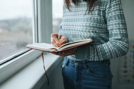 Unrecognizable girl woman stands at window and writes down with pen on paper personal thoughts and...
