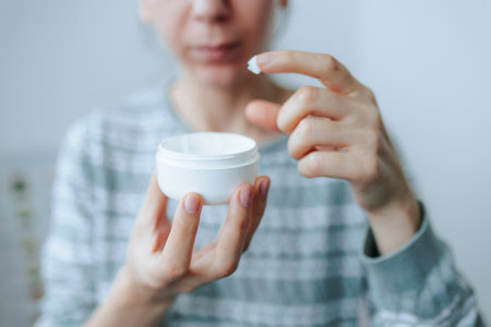 Close up female hands holding jar of face cream rubbing moisturizer applying cream in skin of face. Young woman taking care of beauty. Lady applying seasonal protection cream on face skin.の写真素材