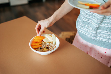 There is a white bowl with oatmeal and fruits on the table, a woman hand beautifully lays out slices of fruit. Delicious healthy breakfast. Healthy food concept. Unrecognizable.の写真素材