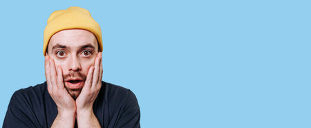 A staring guy wearing a yellow cap, in a studio shot with a blue background, is seen with a...