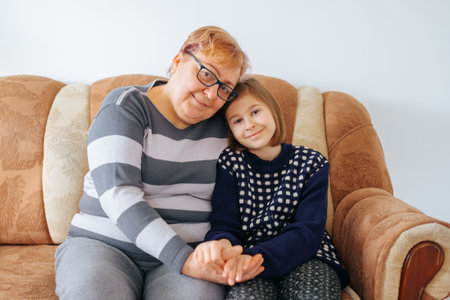 A touching of a grandmother and granddaughter sitting side by side on the couch looking at camera, holding hands and gazing into each others eyes with love and affection.の写真素材