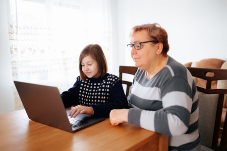 a senior woman is sitting with her grandchild on the chair in their cozy living room. The kid is explaining something they learned on their laptop while the grandmother attentively listens,の写真素材