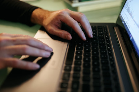 A close-up shot of a pair of hands typing away on a laptop, illuminated by the glow of the screen in a dimly-lit room at nighttime. The laptops top view and touchpad are visible,の写真素材